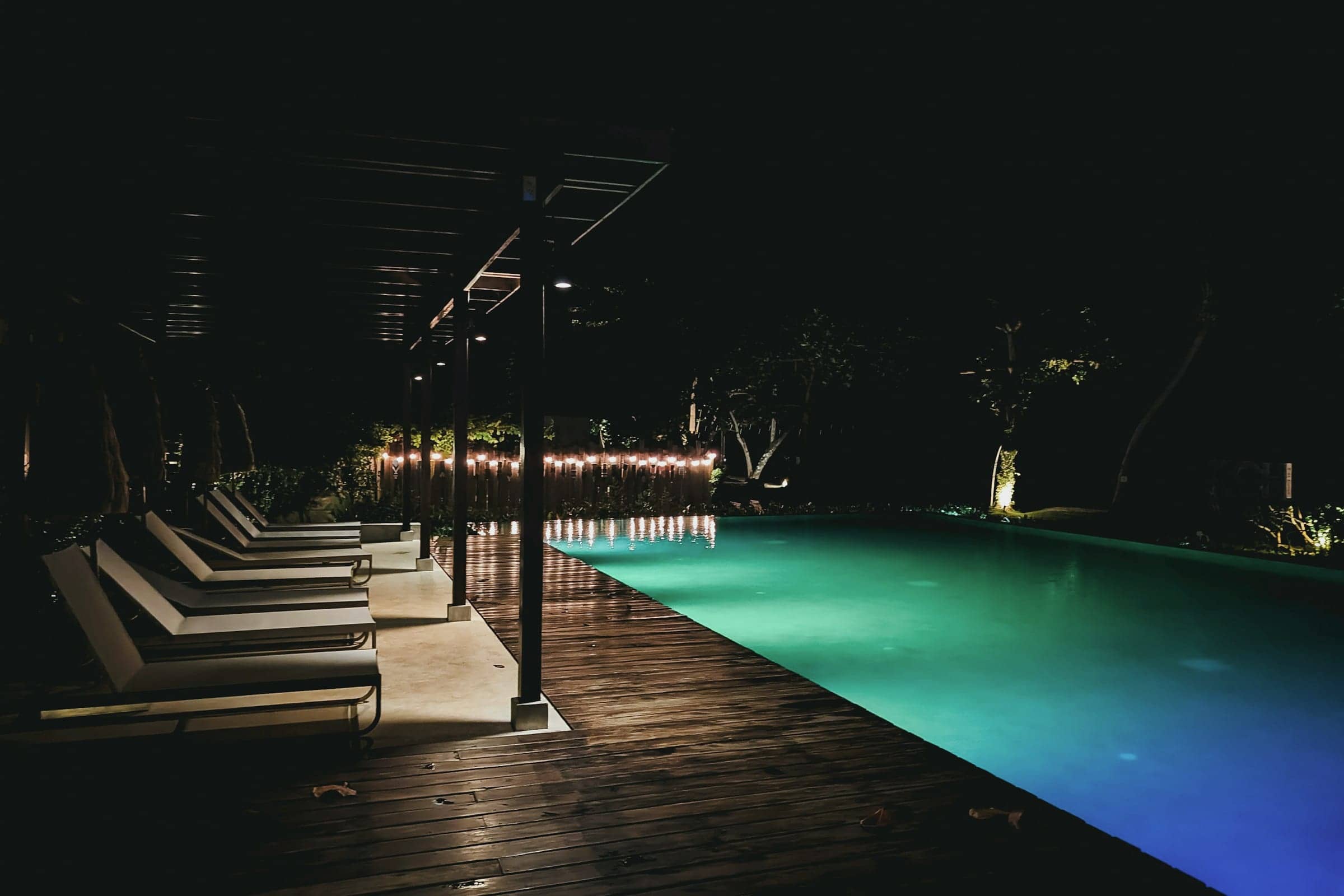 Hotel pool deck at night, with loungers along a lit turquoise pool and string lights under a timber pergola.